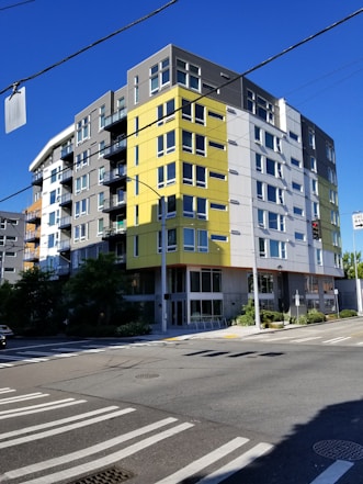 A modern, multi-story apartment building with a mix of yellow, gray, and white panels is situated at the corner of an intersection. The structure features numerous windows and small balconies. The area is clear, with blue skies overhead and a crosswalk visible at the street level.