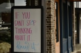 A sandwich board with a motivational message written in colorful letters stands outside a brick building. The text on the board reads 'If you can't stop thinking about it... buy it.' The building features wooden doors and dark blue trim.