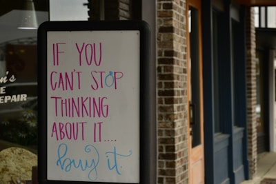 A sandwich board with a motivational message written in colorful letters stands outside a brick building. The text on the board reads 'If you can't stop thinking about it... buy it.' The building features wooden doors and dark blue trim.