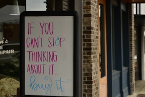 A sandwich board with a motivational message written in colorful letters stands outside a brick building. The text on the board reads 'If you can't stop thinking about it... buy it.' The building features wooden doors and dark blue trim.