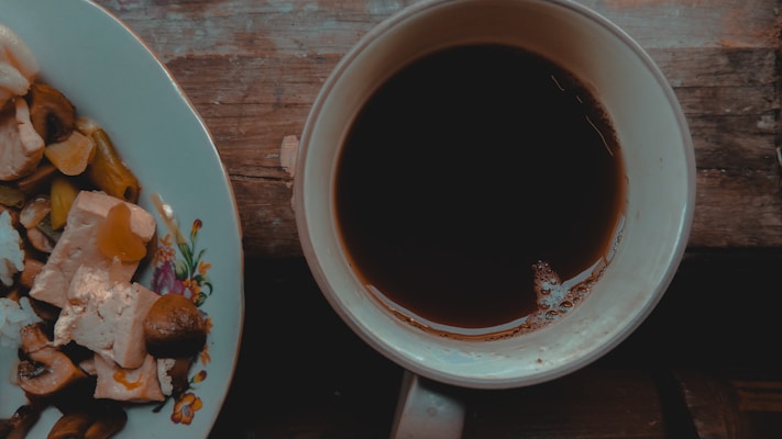 A wooden table holds a white plate with a floral design, containing pieces of tofu, mushrooms, and vegetables like bell peppers. Beside the plate, a white cup filled with dark coffee sits with visible bubbles on its surface.