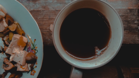 A wooden table holds a white plate with a floral design, containing pieces of tofu, mushrooms, and vegetables like bell peppers. Beside the plate, a white cup filled with dark coffee sits with visible bubbles on its surface.