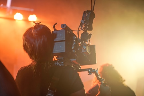 A person is operating a professional video camera amidst a warm, ambient lighting setting. The equipment is mounted on a rig, and the scene appears to be part of a film or video production environment.