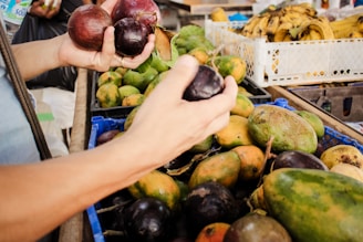 Two hands are holding dark purple passion fruits at a market stall filled with a variety of fresh fruits, including mangoes and bananas. The scene suggests selection and purchasing in a vibrant market setting.