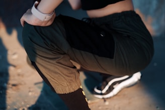 A person is kneeling on the ground, wearing olive green joggers with black accents and black sneakers with white soles. The person's hand rests on their knee, and they are wearing a smartwatch. The lighting creates a play of shadows and highlights on the ground and the clothing.