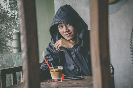 A person wearing a dark hoodie is seated at a wooden table in a dimly lit setting. The person has a relaxed posture and holds a cigarette. In front of them, there’s a glass with a red straw, a phone, and a small red-lidded container. The background contains an outdoor view with blurred greenery.