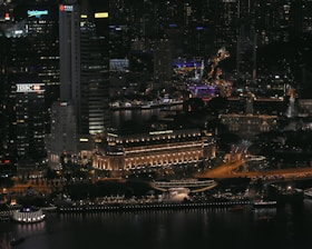 A bustling cityscape at night with illuminated skyscrapers and buildings. Prominent signage includes HSBC and Maybank. The Fullerton Hotel is noticeable, featuring warm lighting. The streets and pathways are well-lit, with visible traffic and activity.