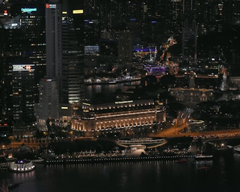 A bustling cityscape at night with illuminated skyscrapers and buildings. Prominent signage includes HSBC and Maybank. The Fullerton Hotel is noticeable, featuring warm lighting. The streets and pathways are well-lit, with visible traffic and activity.