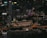 A bustling cityscape at night with illuminated skyscrapers and buildings. Prominent signage includes HSBC and Maybank. The Fullerton Hotel is noticeable, featuring warm lighting. The streets and pathways are well-lit, with visible traffic and activity.