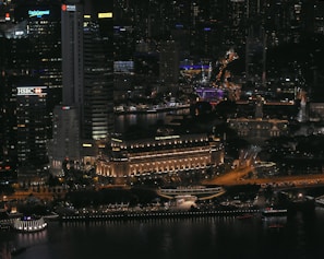 A bustling cityscape at night with illuminated skyscrapers and buildings. Prominent signage includes HSBC and Maybank. The Fullerton Hotel is noticeable, featuring warm lighting. The streets and pathways are well-lit, with visible traffic and activity.