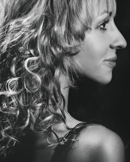 A black and white profile portrait of a person with curly hair. The lighting accentuates the texture of the hair and the contours of the face. The image has a soft focus and a classic, timeless feel.