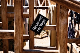 A wooden frame with various textiles hanging over it, featuring a black sign that reads 'Reserved for Yoga.' Sunlight casts shadows, creating a rustic and peaceful atmosphere.