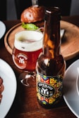 A bottle and a glass of Asperius Hop Trop IPA are placed on a wooden table. The bottle label features a colorful geometric design, and the glass is filled with amber beer topped with a light foam. In the background, there is a plate with a burger and a hint of lettuce peeking out. The setting suggests a casual dining or pub environment.