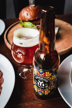 A bottle and a glass of Asperius Hop Trop IPA are placed on a wooden table. The bottle label features a colorful geometric design, and the glass is filled with amber beer topped with a light foam. In the background, there is a plate with a burger and a hint of lettuce peeking out. The setting suggests a casual dining or pub environment.