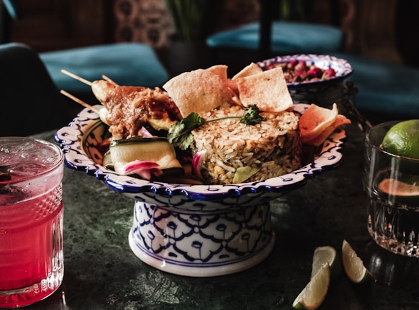 A decorative dish holds a serving of seasoned rice garnished with cilantro, accompanied by skewered grilled meat and crisp crackers. Beside the dish, a vibrant pink drink in a textured glass and a glass containing lime is visible, while lime wedges are placed on the table.