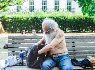 A shirtless elderly man with a long white beard and sunglasses sits on a park bench. He is rummaging through a backpack. Next to him, there is a bottle of drink and some shopping bags. The background features other people sitting on the grass and a hedge with a white building visible behind it.
