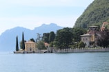 A scenic view of a tranquil lakeside, bordered by elegant villas set against lush greenery and distant mountains. Tall cypress trees and other dense foliage surround the historic-style buildings, creating a serene and picturesque landscape.