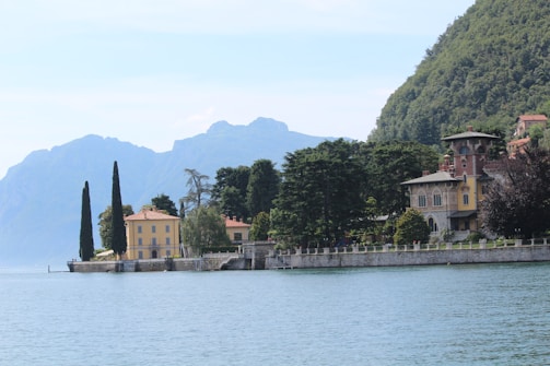 A scenic view of a tranquil lakeside, bordered by elegant villas set against lush greenery and distant mountains. Tall cypress trees and other dense foliage surround the historic-style buildings, creating a serene and picturesque landscape.