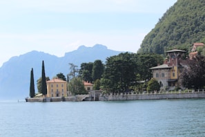 A scenic view of a tranquil lakeside, bordered by elegant villas set against lush greenery and distant mountains. Tall cypress trees and other dense foliage surround the historic-style buildings, creating a serene and picturesque landscape.