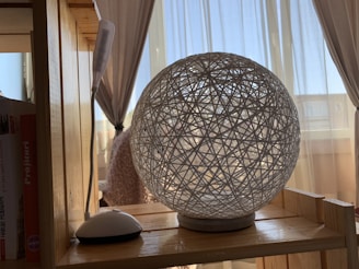 A decorative spherical lamp made of intertwined white strings is placed on a wooden shelf. There is a small table lamp with a flexible neck nearby, and a book is partially visible on the shelf. Curtains and bright natural light suggest a window in the background.