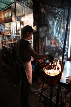 A street food vendor is cooking over an open flame, wearing a white apron and black cap. The scene is set at night, illuminated by nearby artificial lights. The stall has metal structures and signage in another language, with kitchen items and ingredients visible. The environment appears bustling and lively.