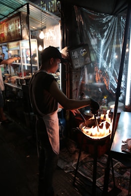 A street food vendor is cooking over an open flame, wearing a white apron and black cap. The scene is set at night, illuminated by nearby artificial lights. The stall has metal structures and signage in another language, with kitchen items and ingredients visible. The environment appears bustling and lively.
