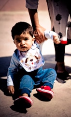 A young child is sitting on the ground while someone holds their arm to assist them. The child is wearing blue overalls, a light-colored shirt, a bib with a cartoon dog design, and bright red shoes. A part of another person's body is visible, wearing dark pants and shoes with a decorative tree pattern on the clothing.