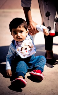 A young child is sitting on the ground while someone holds their arm to assist them. The child is wearing blue overalls, a light-colored shirt, a bib with a cartoon dog design, and bright red shoes. A part of another person's body is visible, wearing dark pants and shoes with a decorative tree pattern on the clothing.