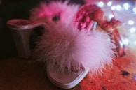 A fluffy pink slipper with a furry texture in the foreground, surrounded by soft, colorful lighting. The background is slightly blurred, adding a dreamy effect to the scene.
