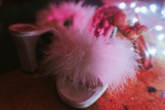 A fluffy pink slipper with a furry texture in the foreground, surrounded by soft, colorful lighting. The background is slightly blurred, adding a dreamy effect to the scene.