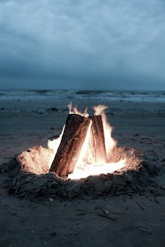 A campfire burns brightly on a sandy beach with the ocean in the background under an overcast sky.