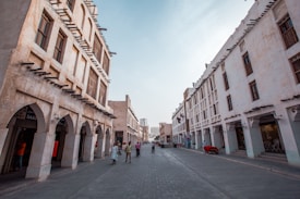 A street view showcasing traditional Middle Eastern architecture with arched windows and wooden beams. The narrow street is flanked by light-colored stone buildings with people leisurely walking, some wearing traditional attire. Flags are visible on the right, and the sky appears clear.