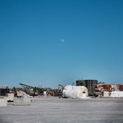 A vast industrial site with scattered debris and large cylindrical tanks lying on their sides. In the background, structures of machinery and equipment are visible, indicating an abandoned or under-construction area. The sky is clear with a visible moon.