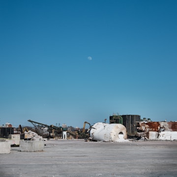 A vast industrial site with scattered debris and large cylindrical tanks lying on their sides. In the background, structures of machinery and equipment are visible, indicating an abandoned or under-construction area. The sky is clear with a visible moon.