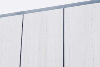 A minimalist exterior wall composed of large vertical white panels, each separated by thin, dark gray lines. The surface appears smooth and uniform, suggesting a modern architectural design.