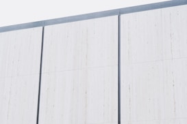 A minimalist exterior wall composed of large vertical white panels, each separated by thin, dark gray lines. The surface appears smooth and uniform, suggesting a modern architectural design.