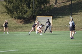 A lacrosse game is taking place on a grassy field, with players wearing uniforms and a referee overseeing the match. A goalie stands in front of a goal with a net, while other players are actively engaged in the game, holding lacrosse sticks.