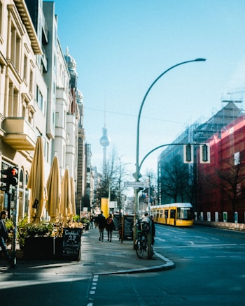 An urban street scene with a mix of modern and traditional architecture, a bright yellow tram, pedestrians walking on the sidewalk, and outdoor cafes with yellow umbrellas. The Fernsehturm tower is visible in the background, and there's a clear blue sky.
