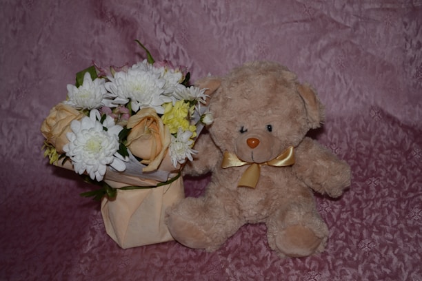 A beige teddy bear with a golden ribbon sits beside a bouquet of flowers wrapped in paper. The bouquet contains white chrysanthemums, yellow and peach roses, and green leaves. Both the teddy bear and flowers are placed on a textured pink background.