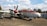 A vintage propeller-driven aircraft is displayed outdoors on a concrete surface. The aircraft features a red and white paint scheme and has sizable windows. The plane is surrounded by trees, a vehicle, and some buildings or structures in the background. A smaller aircraft can be seen flying in the distant sky.