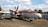 A vintage propeller-driven aircraft is displayed outdoors on a concrete surface. The aircraft features a red and white paint scheme and has sizable windows. The plane is surrounded by trees, a vehicle, and some buildings or structures in the background. A smaller aircraft can be seen flying in the distant sky.