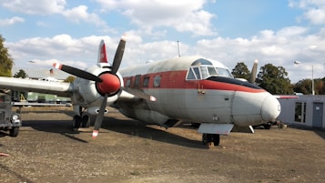 A vintage propeller-driven aircraft is displayed outdoors on a concrete surface. The aircraft features a red and white paint scheme and has sizable windows. The plane is surrounded by trees, a vehicle, and some buildings or structures in the background. A smaller aircraft can be seen flying in the distant sky.