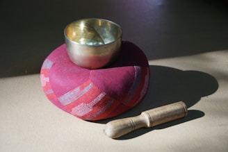 A brass singing bowl is placed on a round red cushion. A wooden mallet is positioned next to the cushion. The scene is lit with natural sunlight, creating soft shadows on the surface.