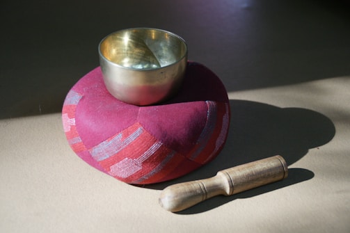 A brass singing bowl is placed on a round red cushion. A wooden mallet is positioned next to the cushion. The scene is lit with natural sunlight, creating soft shadows on the surface.
