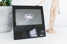 A smart display device with a 'hello' message on the screen next to a wooden artist mannequin. The screen also shows the name 'Jan Antonin Kolar' and the title 'VUI Designer'. A textured plant pot is in the background.