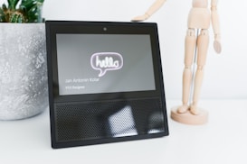A smart display device with a 'hello' message on the screen next to a wooden artist mannequin. The screen also shows the name 'Jan Antonin Kolar' and the title 'VUI Designer'. A textured plant pot is in the background.