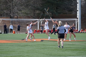 A group of female athletes in athletic gear are playing lacrosse on a grassy field. Several players are competing for the ball in mid-air, with a referee watching the action closely. Spectators are seated in the background, and a soccer goal can be seen behind them. The scene is lively and dynamic, capturing the essence of a competitive sports match.