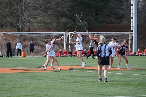 A group of female athletes in athletic gear are playing lacrosse on a grassy field. Several players are competing for the ball in mid-air, with a referee watching the action closely. Spectators are seated in the background, and a soccer goal can be seen behind them. The scene is lively and dynamic, capturing the essence of a competitive sports match.