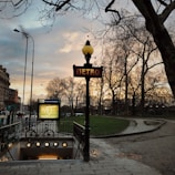 A Paris metro entrance is seen with a traditional art nouveau style sign, under a cloudy sky during sunset. Bare trees line the walkway, and a bicycle is parked near the entrance. The warm light from the streetlamps creates a serene and classic atmosphere.