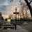 A Paris metro entrance is seen with a traditional art nouveau style sign, under a cloudy sky during sunset. Bare trees line the walkway, and a bicycle is parked near the entrance. The warm light from the streetlamps creates a serene and classic atmosphere.